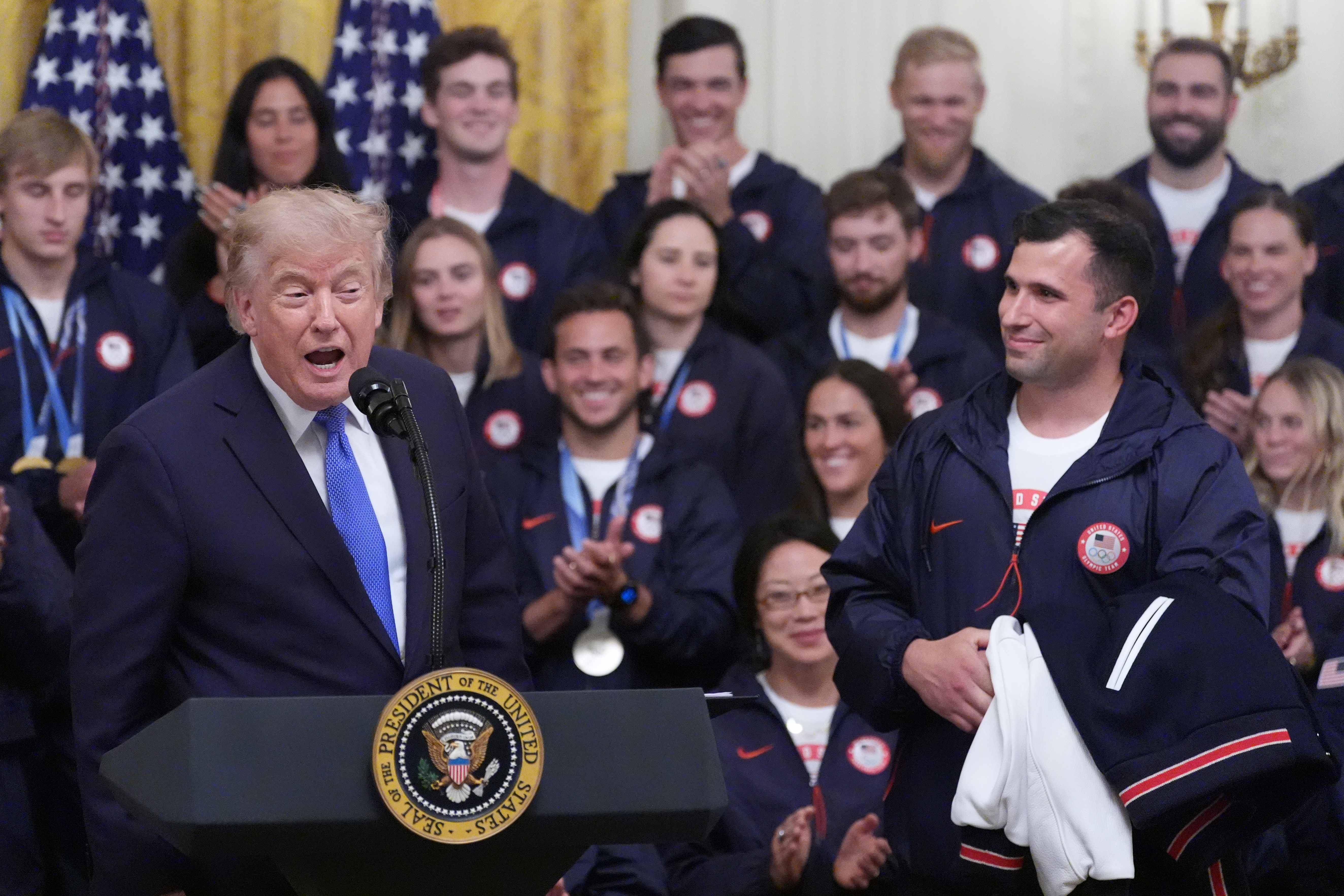 President Donald Trump speaks as bobsledder Frank Del Duca listens during an event for Olympic winter athletes, coaches, staff, and representatives from Team USA in the East Room of the White House, Thursday, Aug. 6, 2026, in Washington. (AP Photo/Alex Brandon)